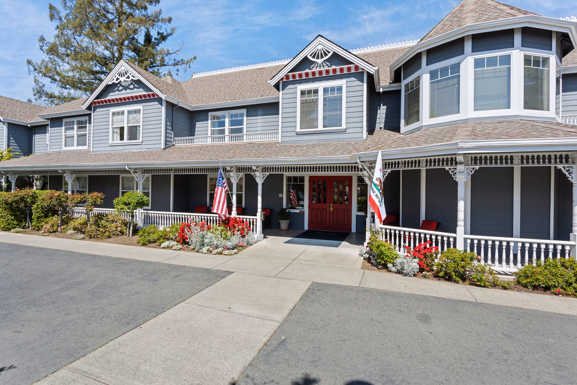 Front exterior of a gray Victorian-style building with a covered porch, red double doors, flags, and flower beds.