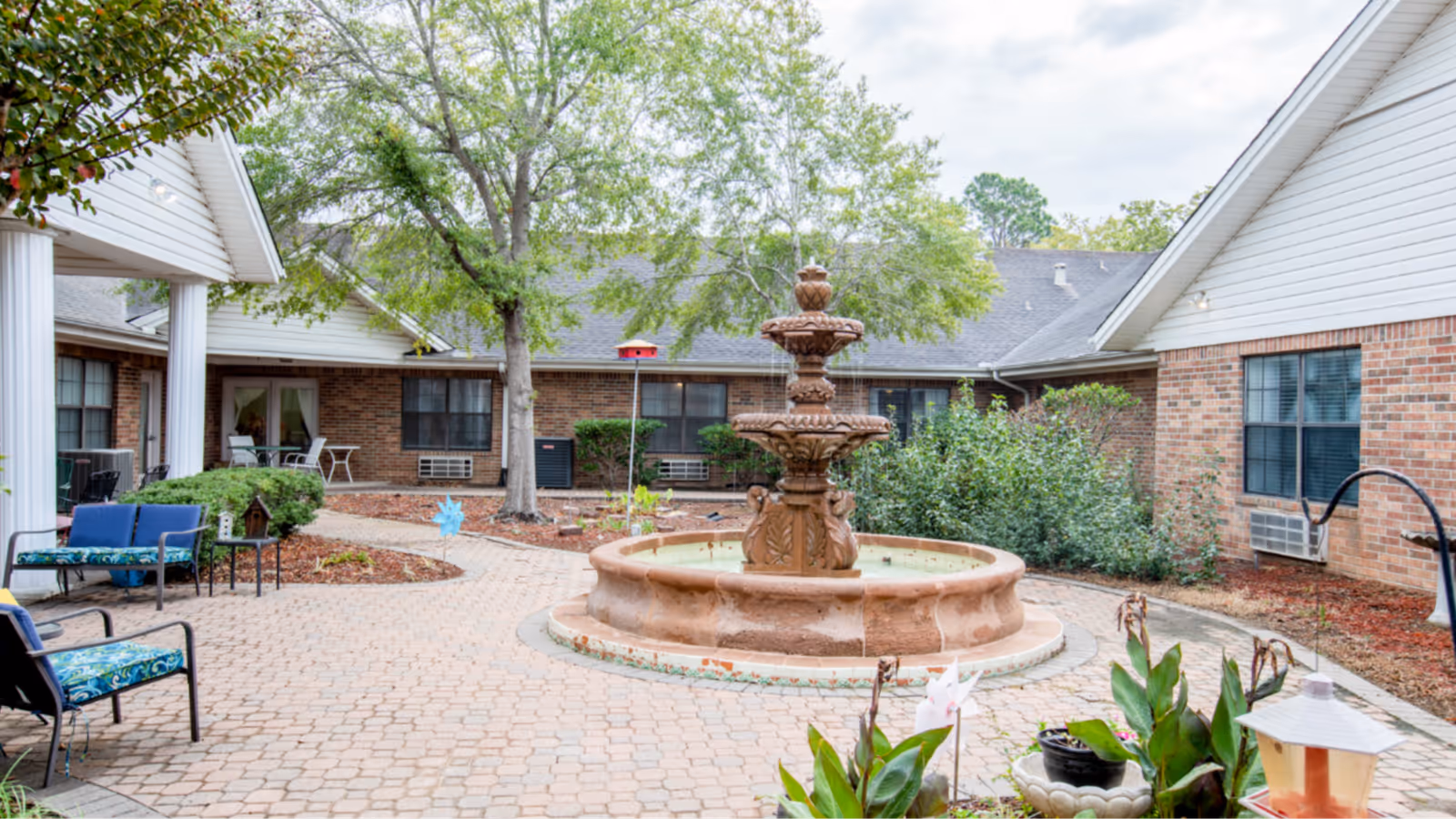 A landscaped courtyard featuring a central tiered stone fountain, patio seating, and surrounding brick assisted-living buildings.