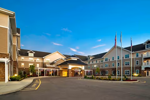 Exterior view of Pinebrook senior living facility at dusk, showing a large multi-story building with a covered entrance, decorated with patriotic bunting, and three flagpoles with flags in front. The sky is clear and blue.