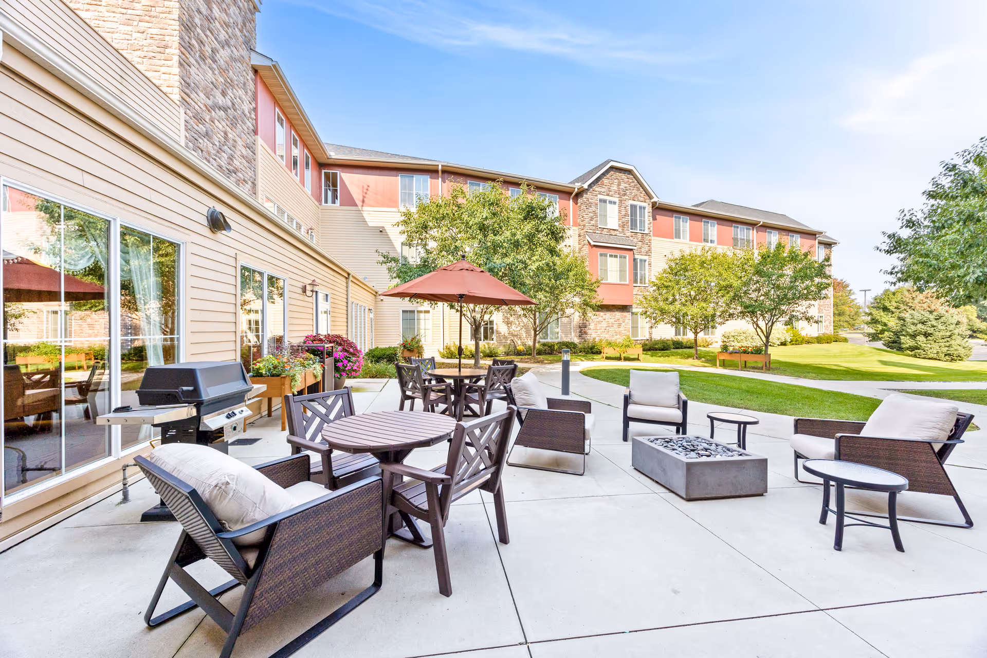 Outdoor patio area at a senior living facility with cushioned chairs, tables, a fire pit, a barbecue grill, and an umbrella. The patio is surrounded by green grass, trees, and a multi-story building with beige and brick exterior walls under a clear blue sky.
