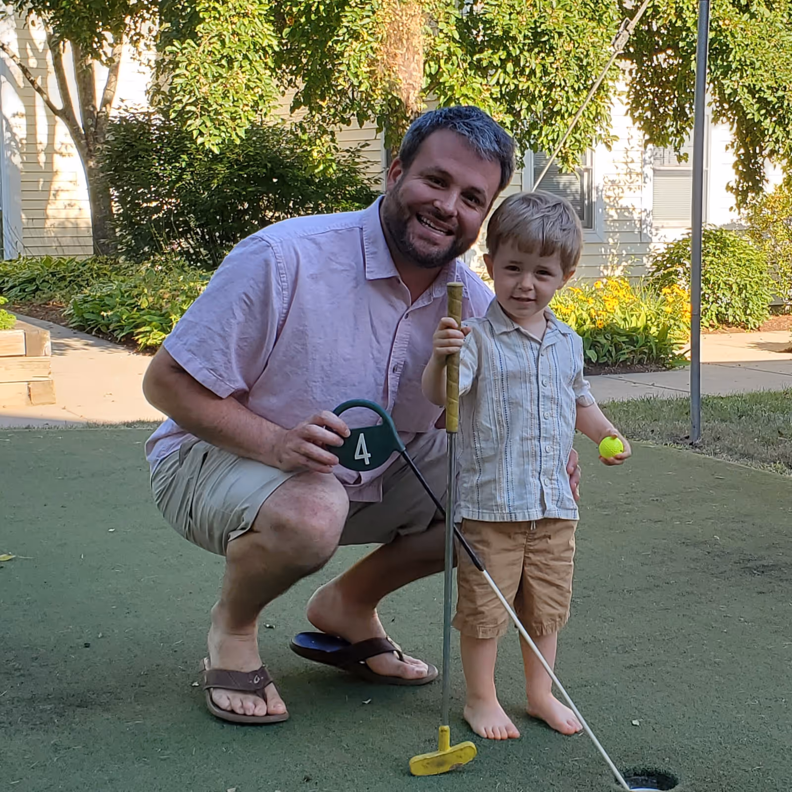 A man and a young boy playing mini golf outdoors. The man is crouching and smiling, holding a mini golf club and a sign with the number 4. The boy is standing barefoot, holding a mini golf club and a yellow golf ball. They are on a green putting surface with trees and bushes in the background.