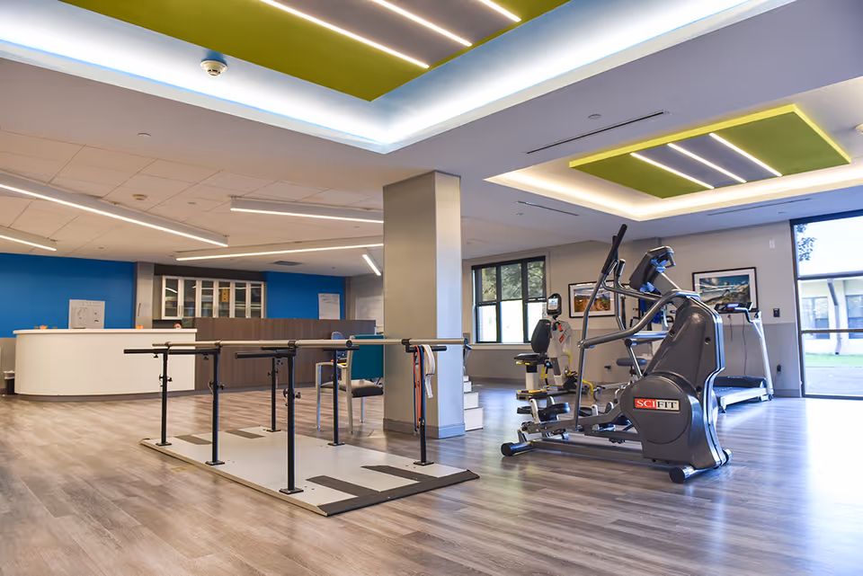 A spacious rehabilitation room with exercise equipment including parallel bars for walking practice and a SciFit elliptical machine. The room has wood-patterned flooring, a blue accent wall with cabinets, and large windows letting in natural light. The ceiling features modern recessed lighting with green and white panels.