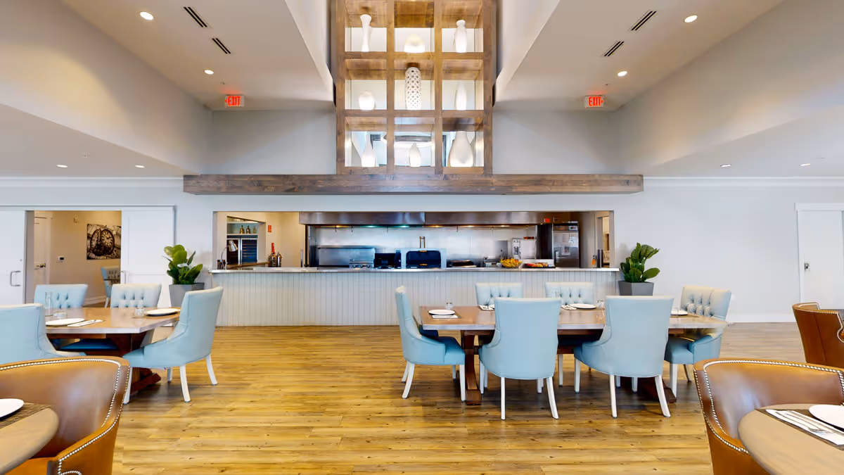 A spacious dining room with wooden floors and light blue upholstered chairs around wooden tables. The room features a large open kitchen area with stainless steel appliances and a decorative wooden shelving unit with white vases above the kitchen counter. There are plants on either side of the kitchen area and recessed lighting in the ceiling.