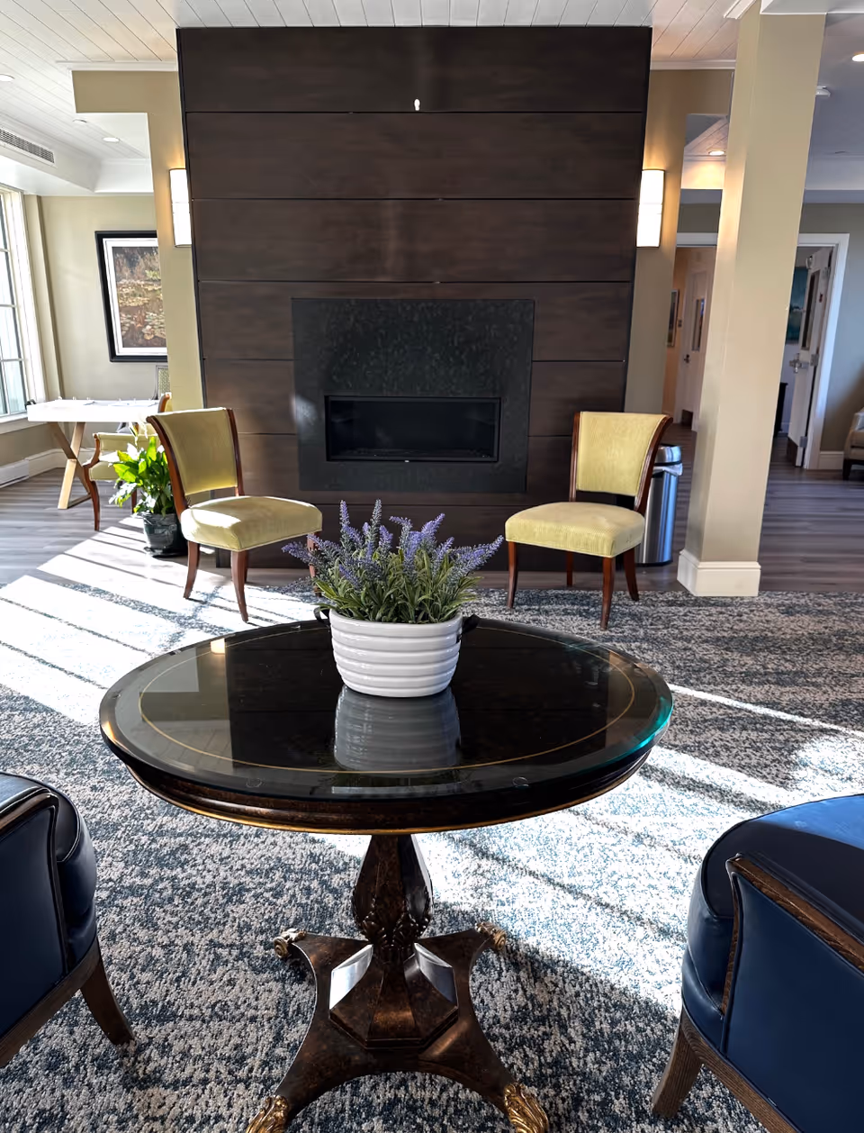 A cozy seating area in a senior living facility featuring a round wooden table with a white pot of purple flowers on top, surrounded by two yellow upholstered chairs and two blue armchairs. Behind the chairs is a modern dark wood-paneled fireplace. The room has large windows letting in natural light, a patterned carpet, and light-colored walls with wall sconces.