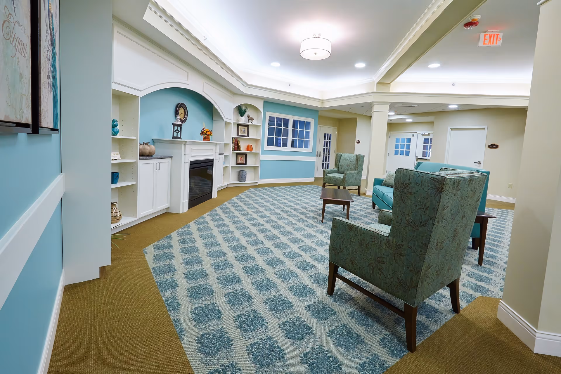 A well-lit senior living facility common area with teal and beige patterned carpet, teal upholstered armchairs, a small wooden coffee table, built-in white shelves with decorative items, and a white fireplace. The walls are painted light blue and beige, with white trim and crown molding. There are framed artworks on the left wall and an exit sign above a door in the background.