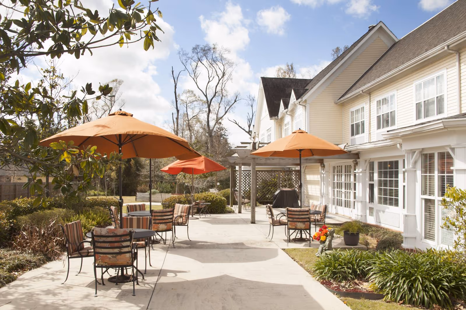 Outdoor patio with tables, striped chairs and orange umbrellas beside the light-colored exterior of a senior living building.