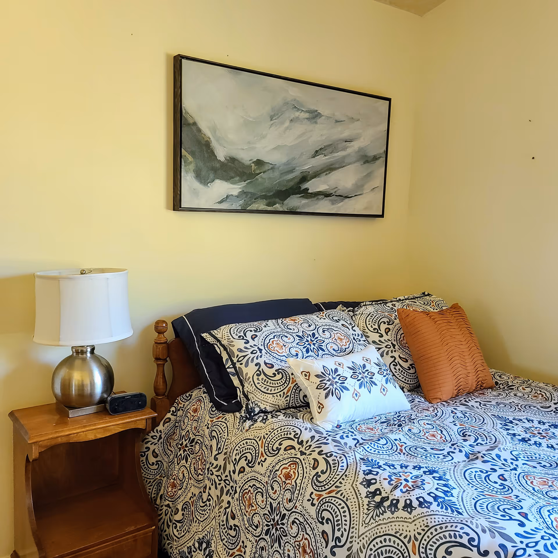A bedroom with a bed covered in a patterned bedspread with blue, white, and orange designs. There are multiple pillows on the bed, including a solid orange one. A wooden nightstand beside the bed holds a silver lamp with a white lampshade and a small black alarm clock. Above the bed hangs a framed abstract painting with shades of gray and white.