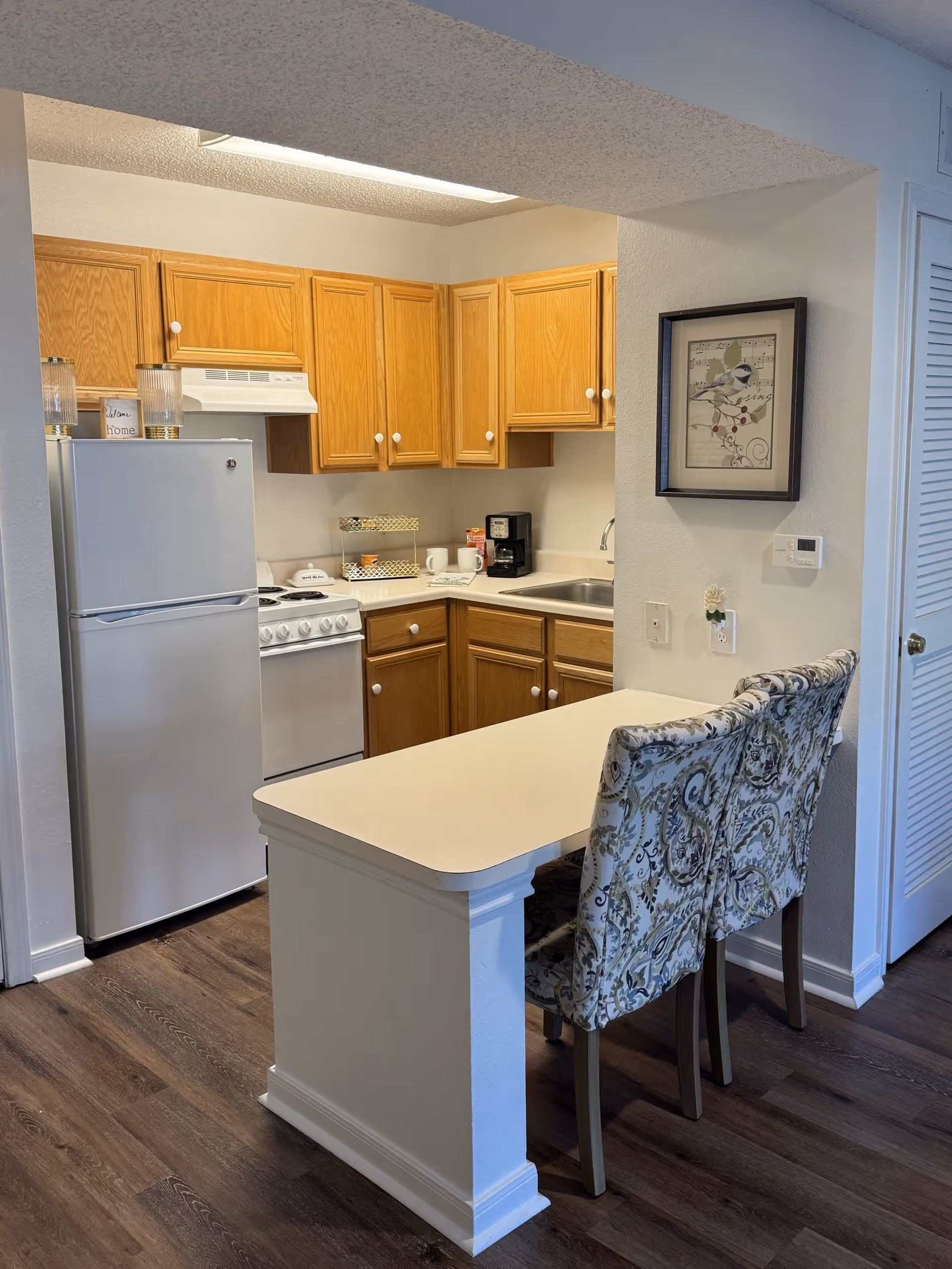 A small kitchen area with wooden cabinets, a white refrigerator, a white stove with an overhead vent, and a countertop with a coffee maker and some cups. In front of the kitchen is a white breakfast bar with two patterned upholstered chairs. A framed picture of a bird hangs on the wall next to a thermostat.
