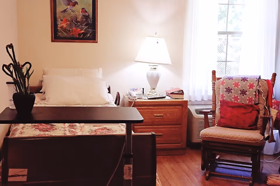 A cozy senior bedroom with a bed, over-bed table, nightstand with a lamp and books, and a cushioned rocking chair by a window.
