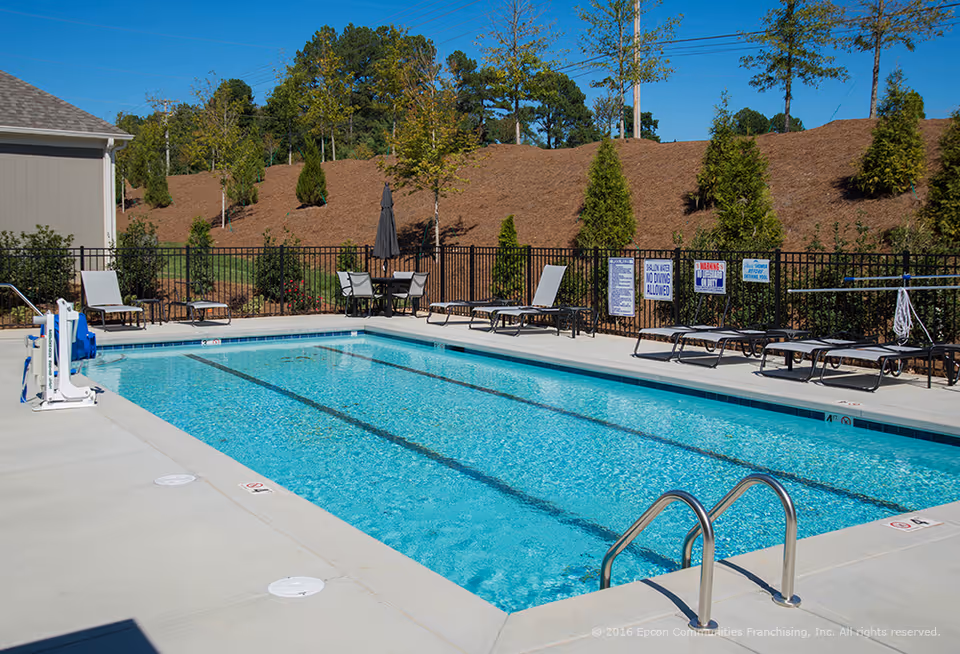 Outdoor swimming pool with clear blue water surrounded by a concrete deck. Several lounge chairs and a table with an umbrella are placed around the pool. There is a black metal fence enclosing the pool area, with trees and shrubs planted on the other side of the fence. The sky is clear and blue.