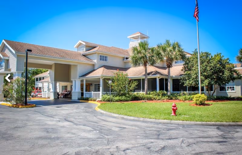 Exterior view of a senior living facility building with a covered entrance, palm trees, a flagpole with an American flag, and well-maintained landscaping including green grass and shrubs under a clear blue sky.