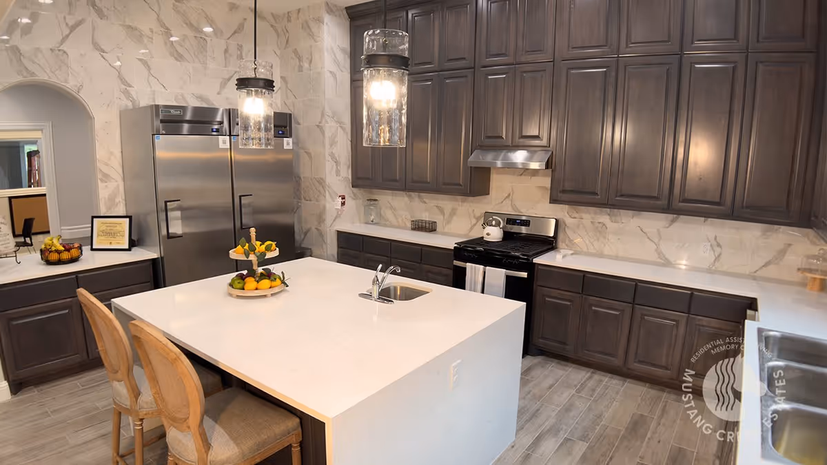 Modern kitchen with dark wood cabinets, a large white island with a built-in sink, two wooden chairs, stainless steel refrigerator and stove, pendant lights, and a fruit stand on the island.