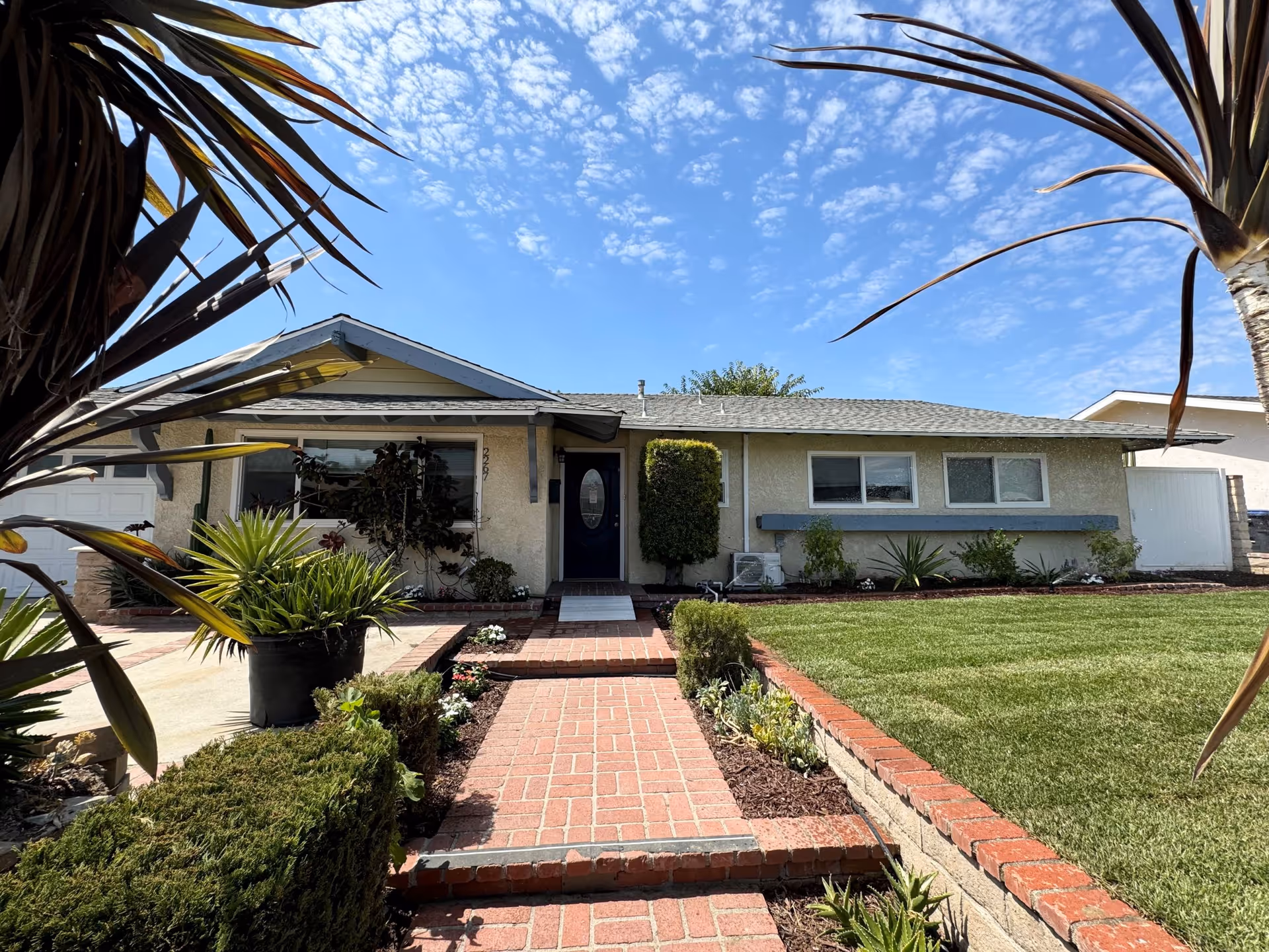 Single-story house with a brick walkway, manicured lawn and tropical plants leading to a blue front door under a sunny sky.