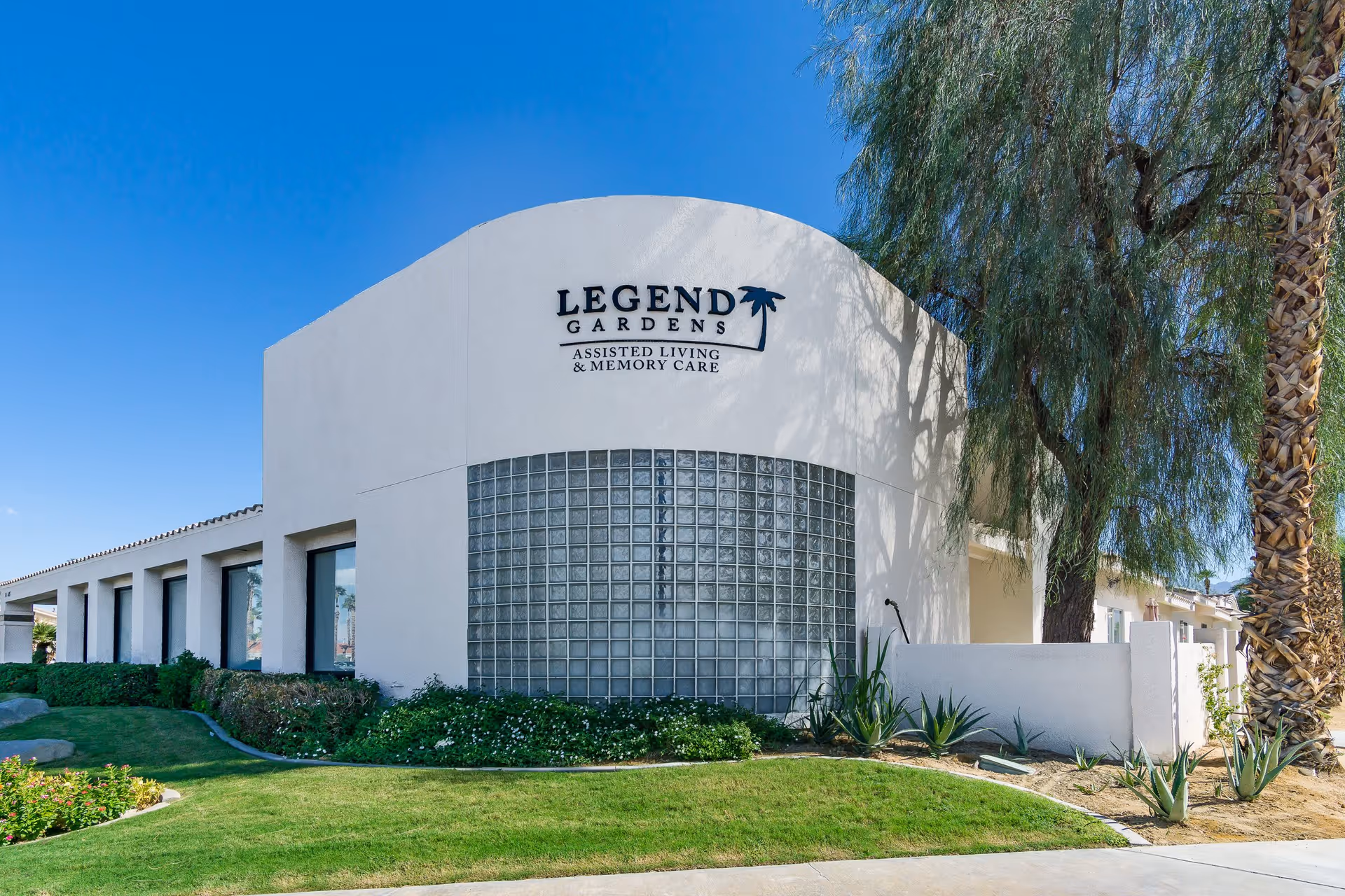 Exterior view of Legend Gardens assisted living and memory care facility with white stucco walls, a curved glass block window, green landscaping, palm trees, and a clear blue sky.
