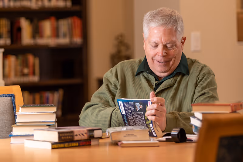 An elderly man with gray hair wearing a green sweater is sitting at a table in a library or reading room, smiling while holding and looking at a book. Several stacks of books are on the table in front of him, and bookshelves filled with books are visible in the background.
