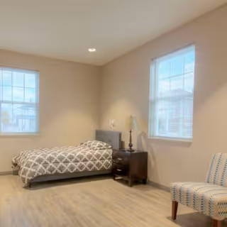 A simple bedroom with a single bed covered in a gray and white patterned bedspread, a dark wooden nightstand with a lamp, two windows letting in natural light, and a striped upholstered chair on the right side.