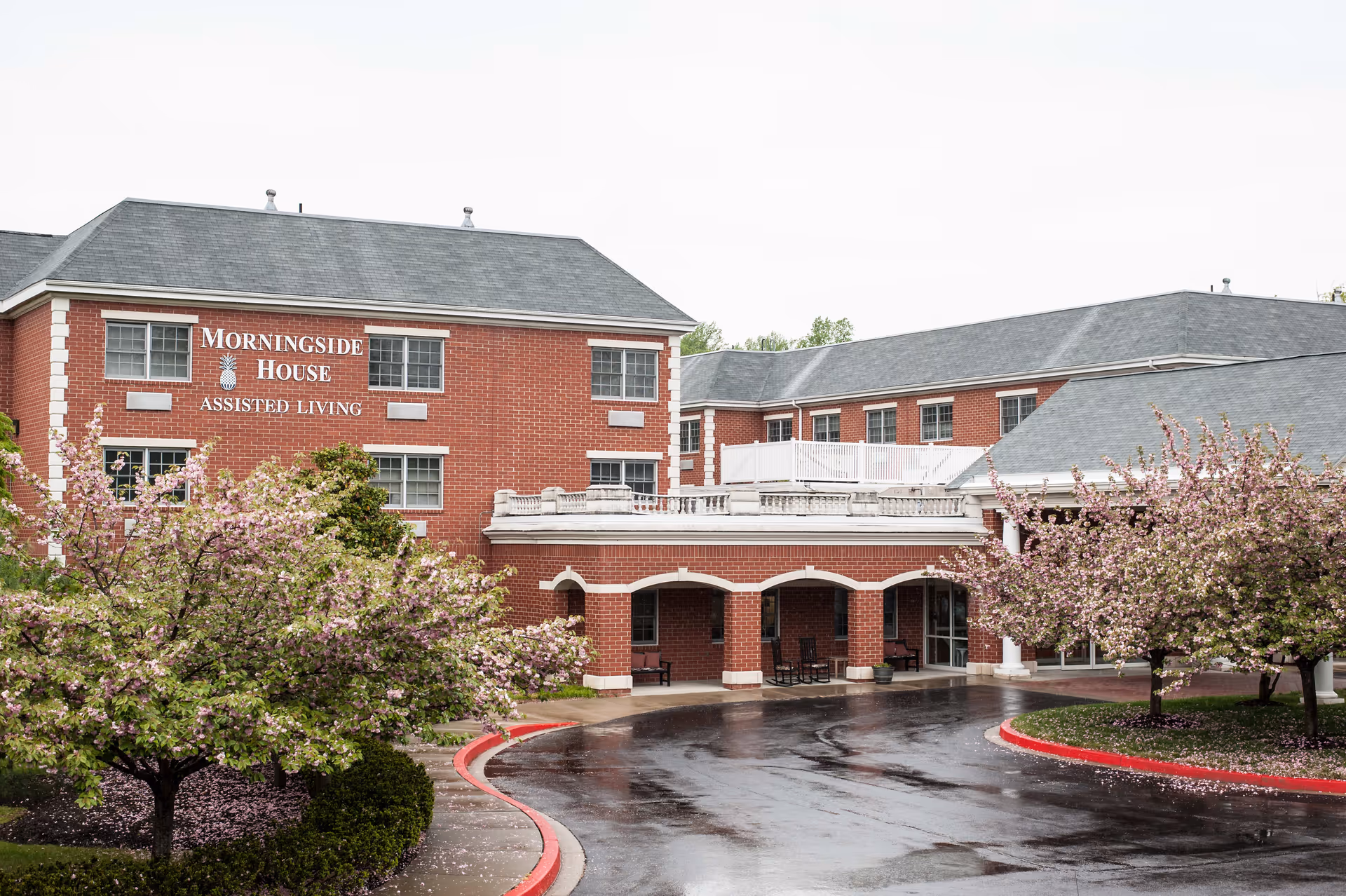Exterior view of Morningside House Assisted Living facility in Ellicott City, showing a red brick building with multiple windows, a covered entrance with columns, and blooming trees with pink flowers in the foreground on a wet driveway.