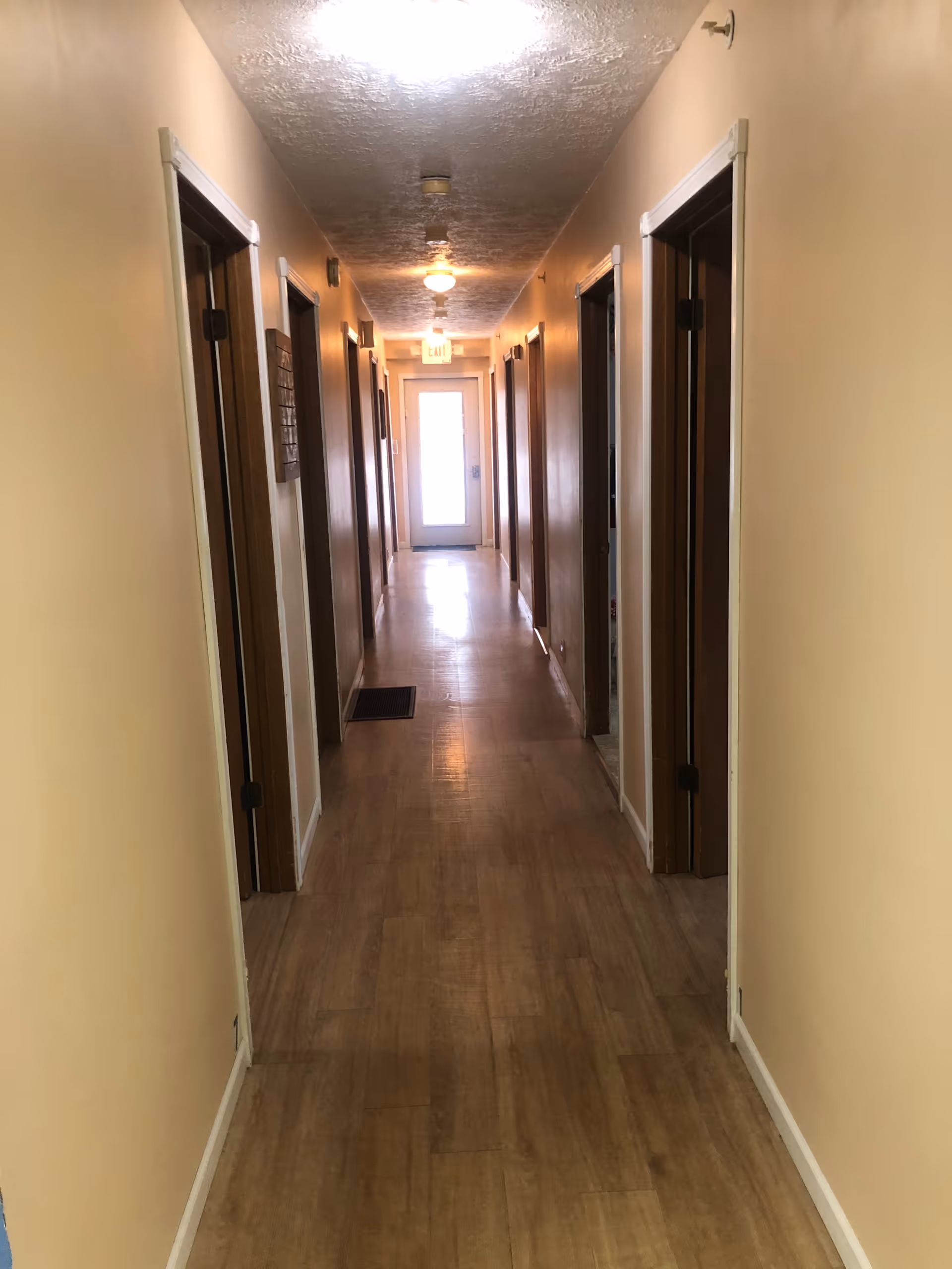 A long indoor hallway with wooden flooring and beige walls, featuring multiple doorways on both sides and a door with a window at the far end. Ceiling lights illuminate the corridor.