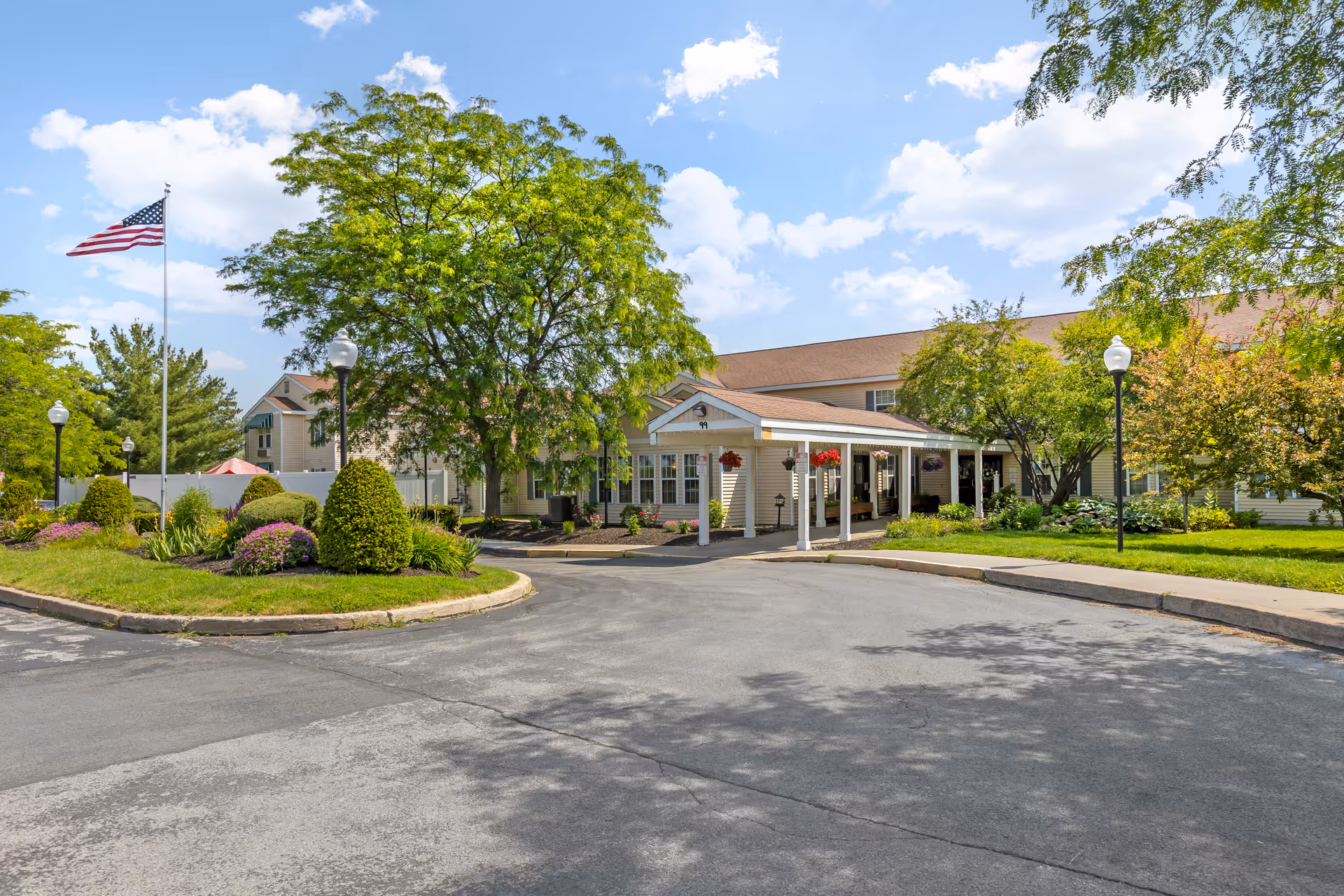 Exterior view of Brookdale Clinton senior living facility showing a driveway entrance with a covered drop-off area, well-maintained landscaping including trimmed bushes and trees, an American flag on a flagpole, and a clear blue sky with some clouds.