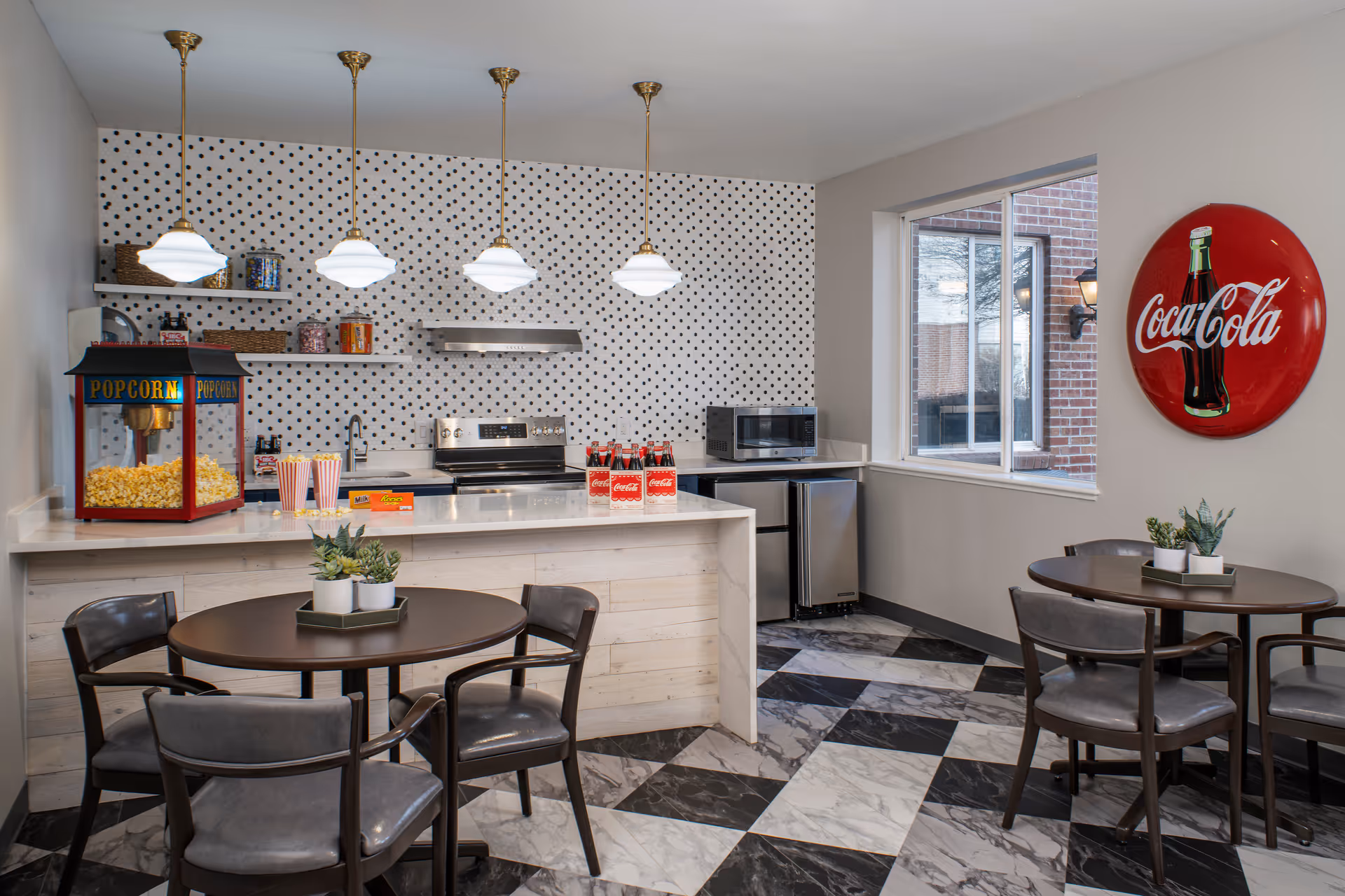 A bright kitchen and dining area with a black and white checkered floor, two round tables with chairs, a popcorn machine on the counter, a stove with a stainless steel hood, a microwave, and a Coca-Cola wall decoration. The kitchen has white countertops and a polka dot backsplash.