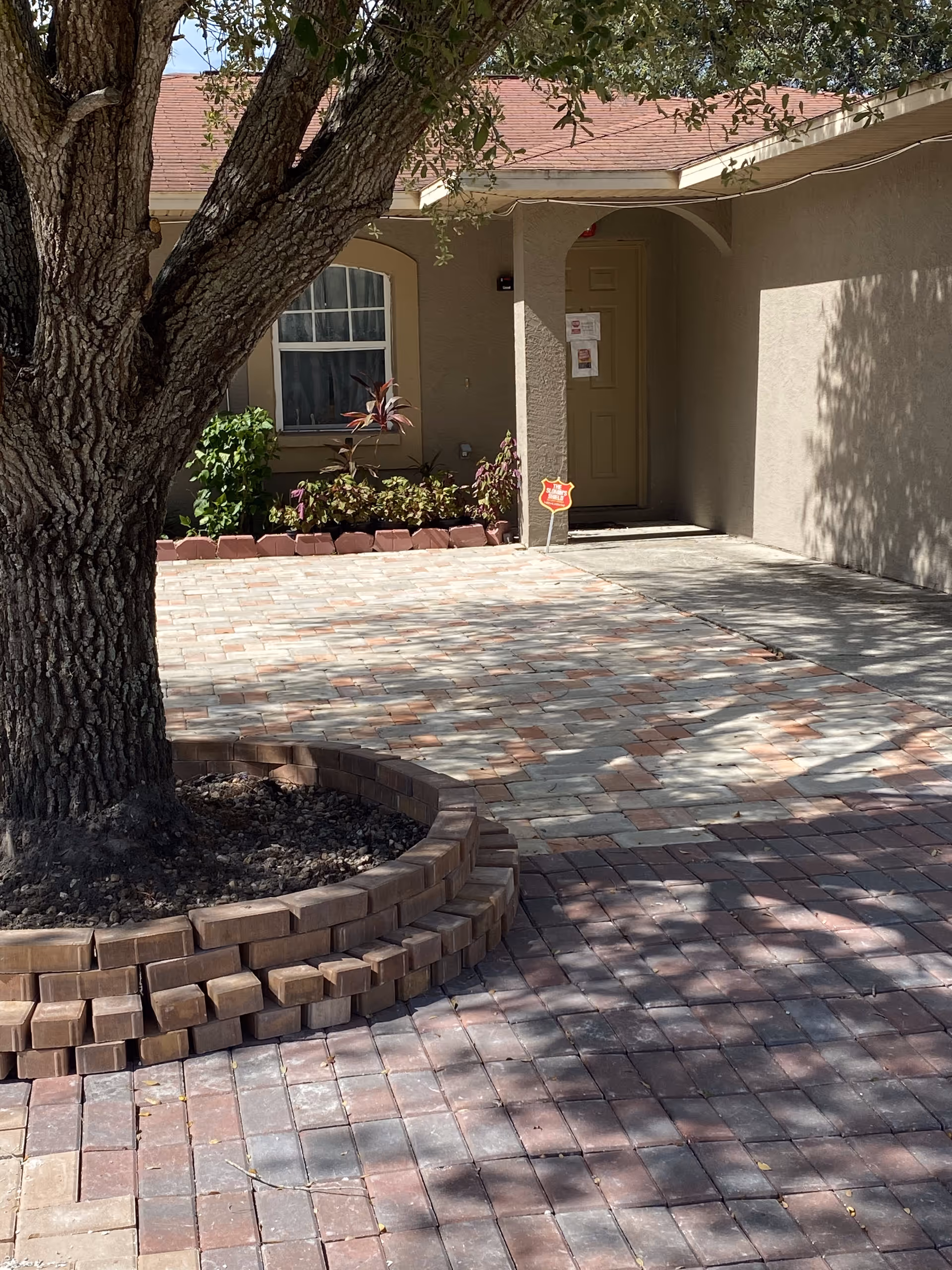 Outdoor view of a facility entrance with a large tree surrounded by a circular brick planter in the foreground. The entrance has a paved walkway and a beige door with some notices posted on it. There are plants and a window to the left of the door, and the building has a light brown exterior with a red roof.