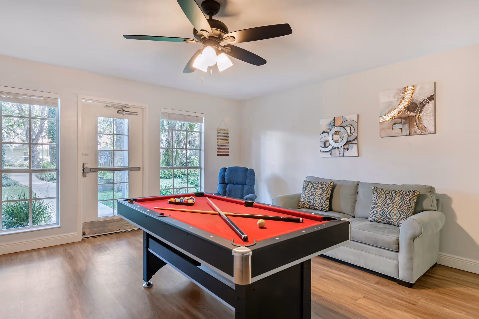 Bright lounge/game room with a red pool table, sofa, recliner, ceiling fan, and large windows and door to the outside.