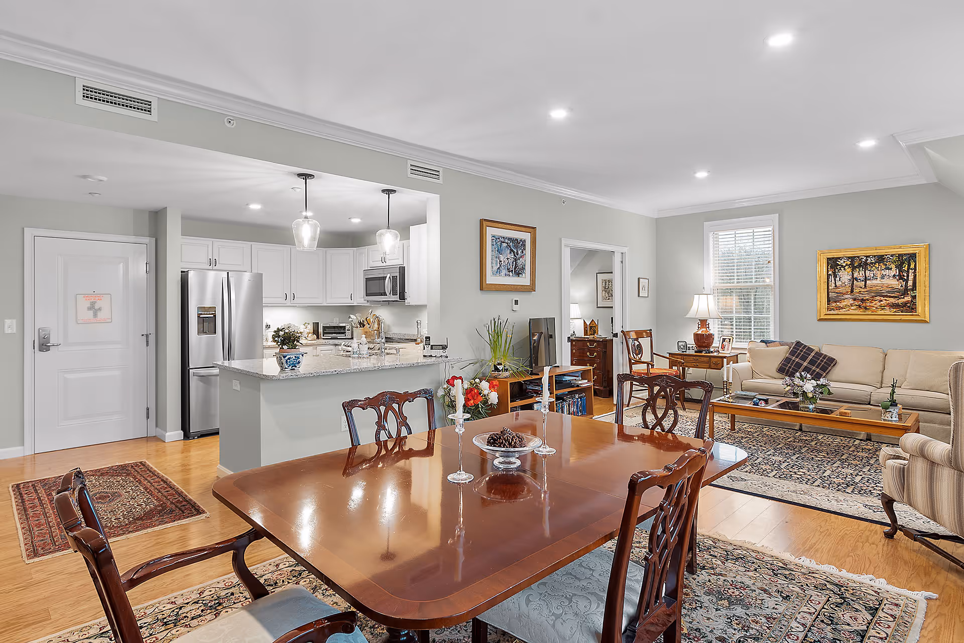 A bright and spacious open-concept living and dining area in a senior living facility. The foreground features a polished wooden dining table with four chairs and decorative candle holders. Behind the dining area is a modern kitchen with white cabinetry, stainless steel refrigerator, and two pendant lights hanging over a granite countertop island. The living room area includes a beige sofa, armchair, wooden coffee table, and two framed paintings on the walls. The room has hardwood floors and several area rugs, with a window letting in natural light.