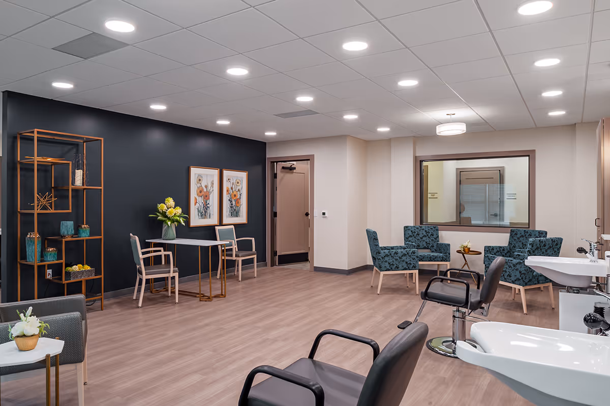 A modern salon area in a senior living facility with two black salon chairs and white sinks in the foreground. The room features wood flooring, a black accent wall with two floral paintings, a small table with two chairs, a decorative shelving unit with vases, and a seating area with four patterned armchairs around a small round table. The ceiling has recessed lighting and a hanging light fixture, and there is a large window looking into another room.