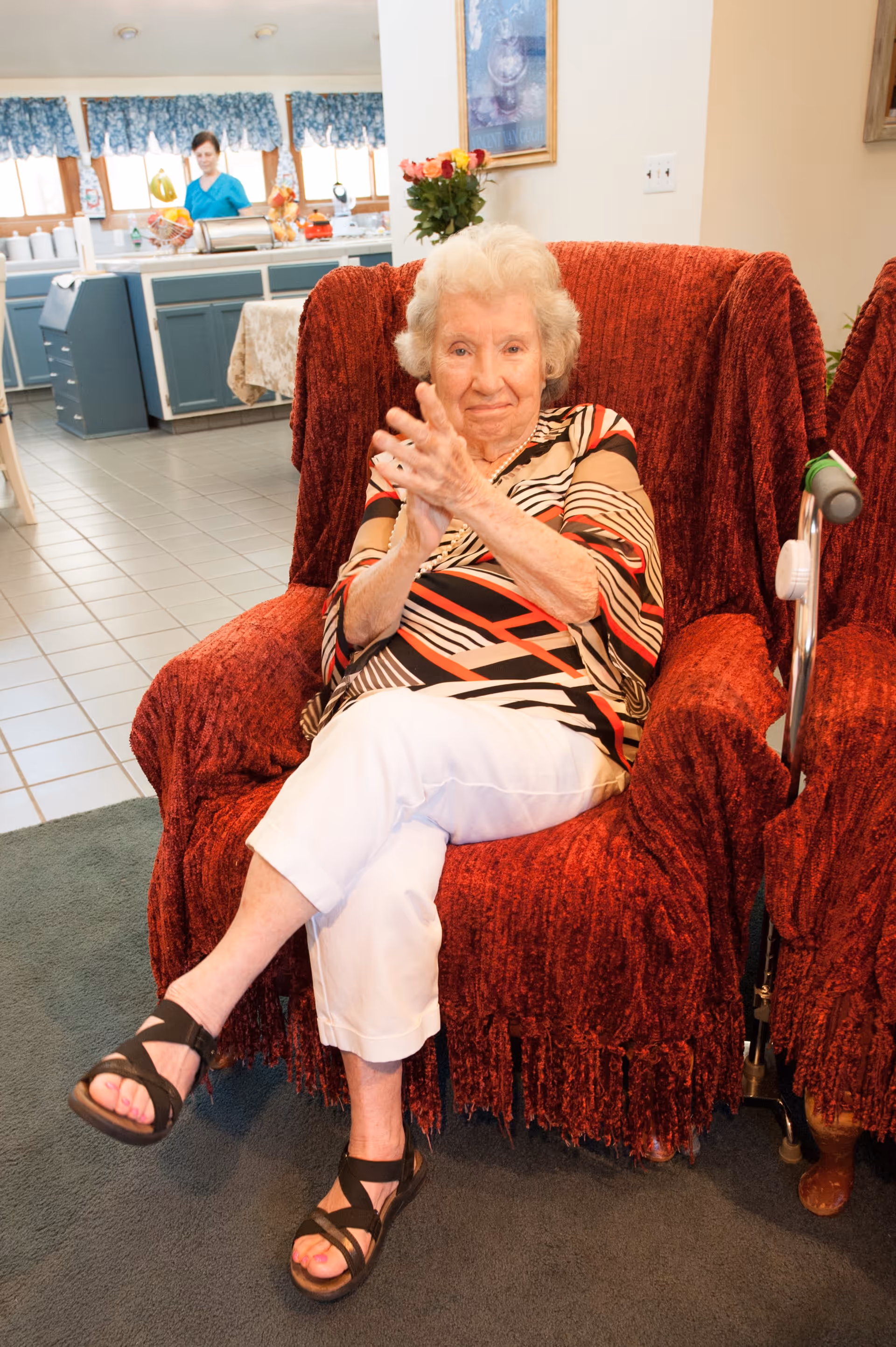 An elderly woman with white hair sitting on a red armchair in a living room area, clapping her hands. She is wearing a patterned top and white pants. In the background, a woman in a blue shirt is seen standing in a kitchen area with blue cabinets and a tiled floor.