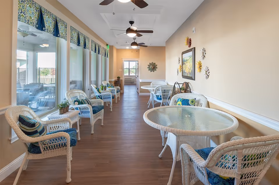 A bright and airy indoor seating area with white wicker chairs and glass-top tables arranged along a hallway. Large windows on one side let in natural light, and the walls are decorated with colorful floral artwork and wall hangings. Ceiling fans with lights hang from the white ceiling, and the floor is a warm wood tone.