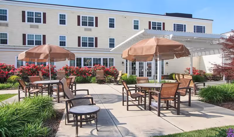 Outdoor patio area at Renaissance Home Pinebrook with several round tables, brown umbrellas, and chairs arranged on a concrete surface. The patio is surrounded by green plants and red flowering bushes, with a three-story building in the background under a clear blue sky.