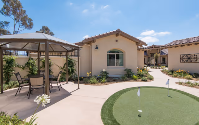 Outdoor courtyard area at ActivCare 4S Ranch featuring a small putting green with two flags, a covered seating area with chairs and a table, surrounded by beige stucco buildings with tiled roofs and landscaped plants under a clear blue sky.