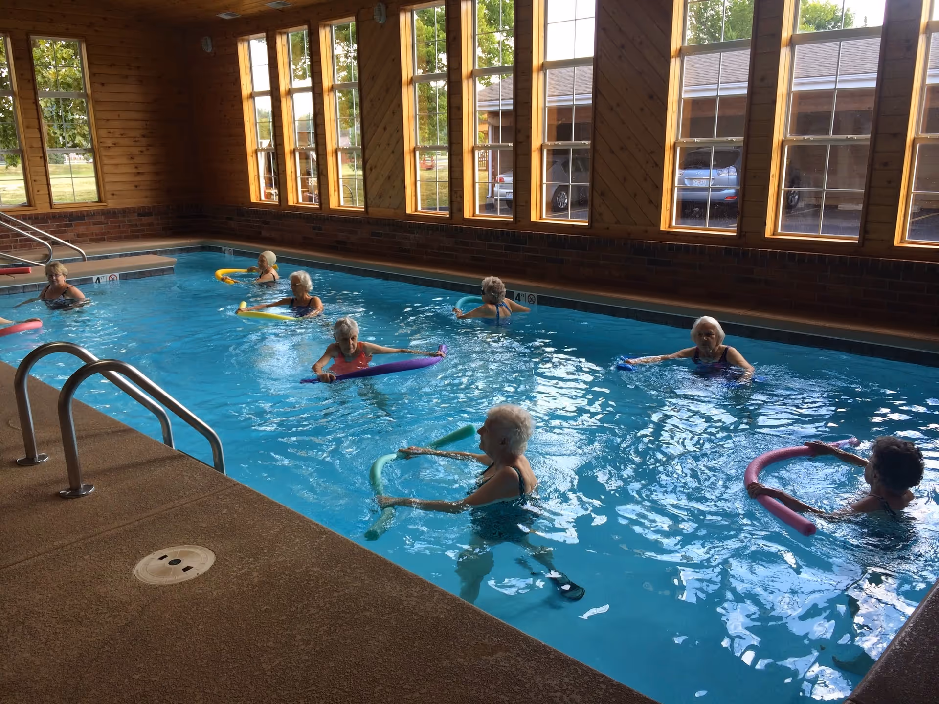 A group of elderly women participating in a water exercise class in an indoor swimming pool with large windows letting in natural light.