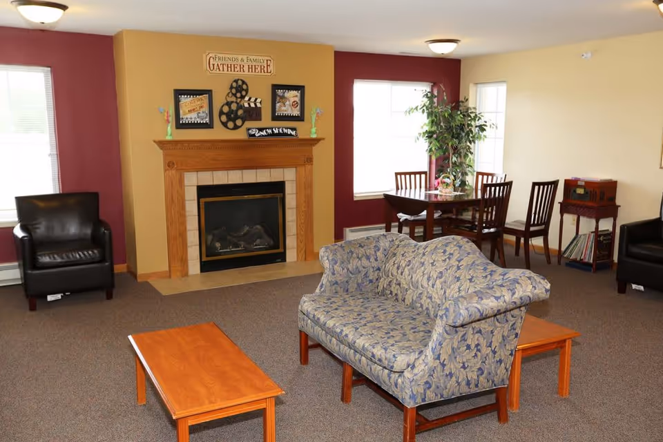 A cozy living room area with a patterned loveseat, two black leather armchairs, a wooden coffee table, and a fireplace with decorative movie-themed wall art above it. There is a dining table with four chairs near two windows, and a potted plant beside the table. The walls are painted in beige and burgundy tones.