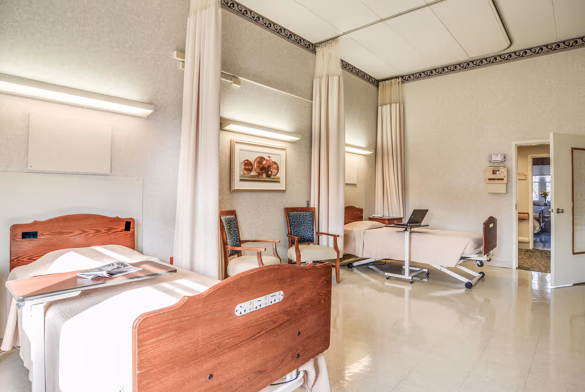 A bright and clean nursing facility room with two hospital beds separated by privacy curtains. Each bed has a bedside table, and there are two upholstered chairs placed between the beds. The room has light-colored walls with a decorative border near the ceiling, a framed picture on the wall, and a door leading to another room.
