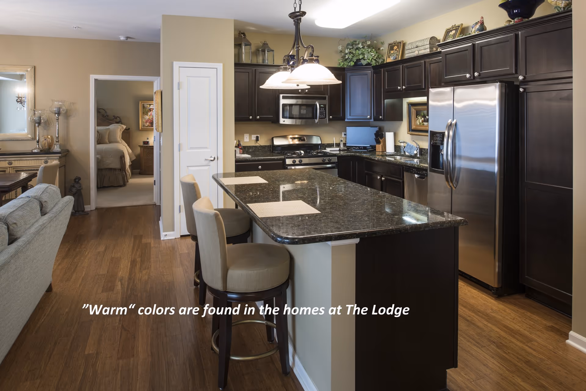 Interior view of a kitchen with dark wood cabinets, stainless steel appliances including a refrigerator and microwave, and a large granite island with two beige cushioned bar stools. The kitchen opens into a living area with a gray sofa and a dining table. A bedroom is visible through an open doorway in the background. Warm beige walls and wooden flooring are present throughout.
