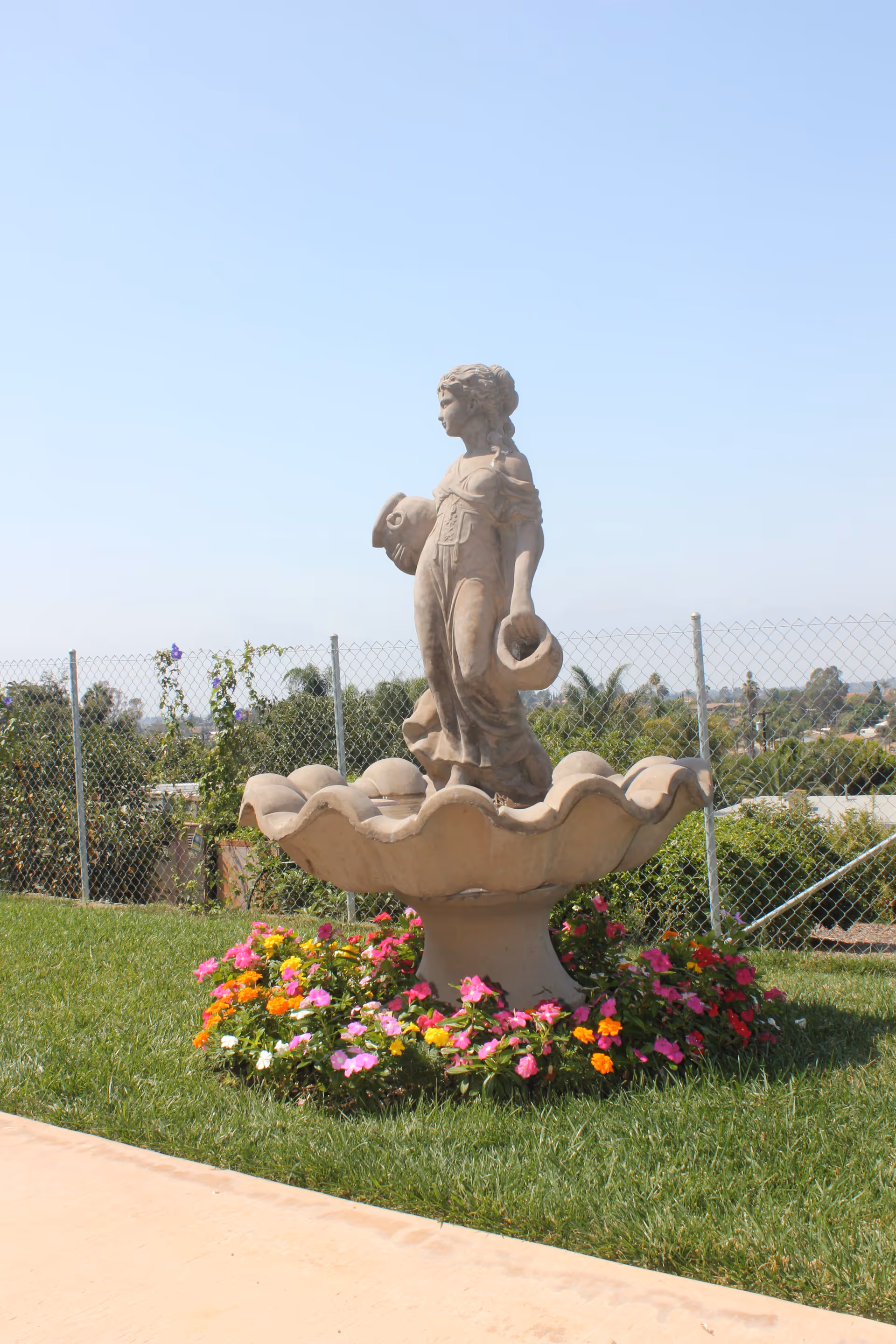 A stone statue of a woman holding two jugs, standing in the center of a large scalloped basin surrounded by colorful flowers on a grassy lawn, with a chain-link fence and trees in the background under a clear blue sky.