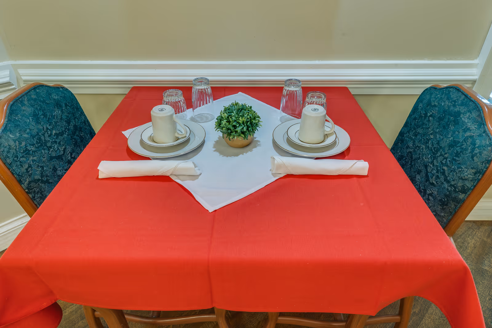A small dining table set for two with a red tablecloth and a white square centerpiece cloth. Each place setting includes a white cup on a saucer, a plate, a rolled white napkin, and an upside-down glass. A small green potted plant is placed in the center of the table. Two green upholstered chairs with wooden frames are positioned on either side of the table.