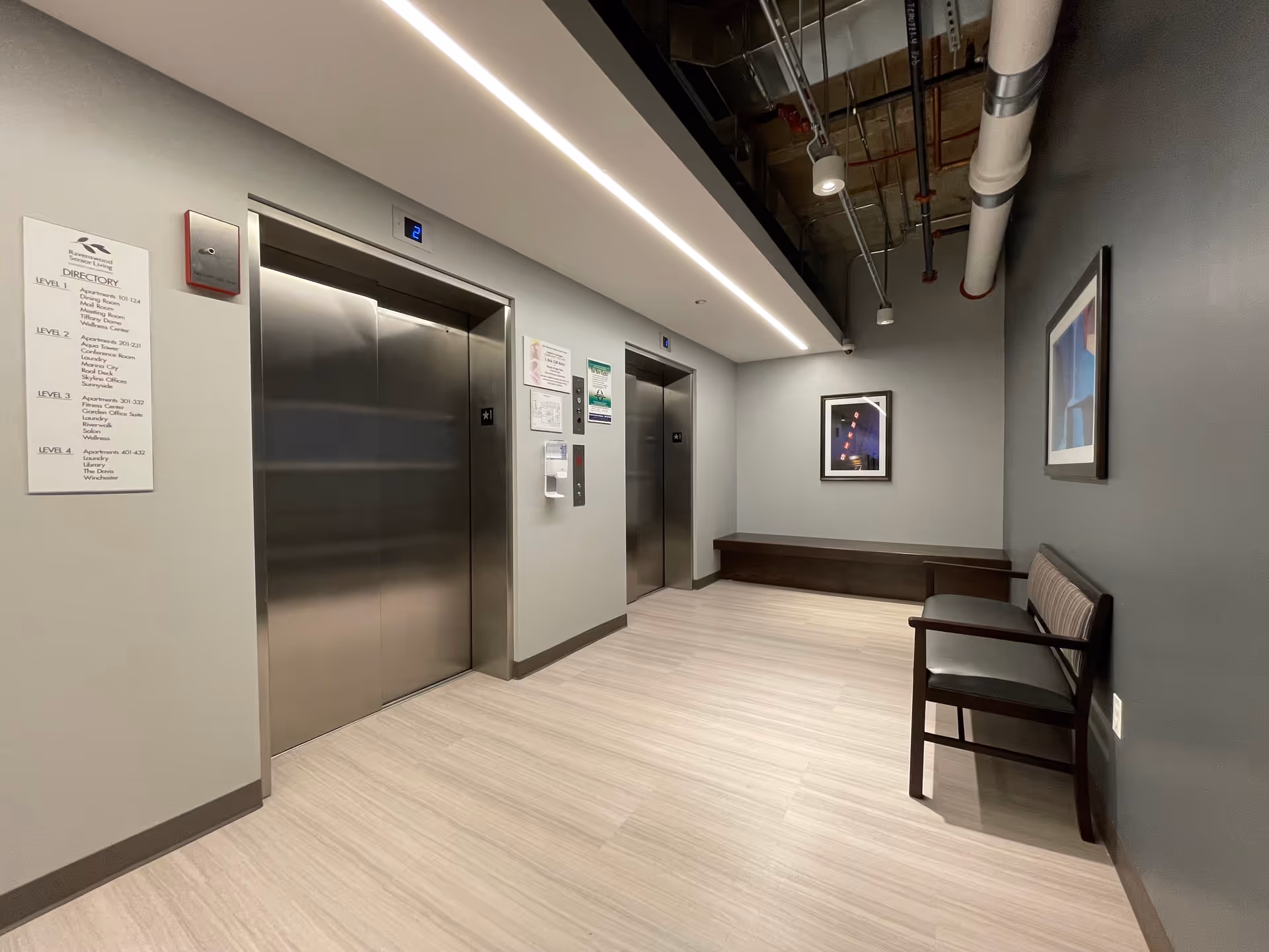 Interior view of an elevator lobby area in Ravenswood Supportive Living facility featuring two closed stainless steel elevator doors, a directory sign on the left wall, a bench along the right wall, two framed abstract artworks, and exposed ceiling pipes with modern lighting.
