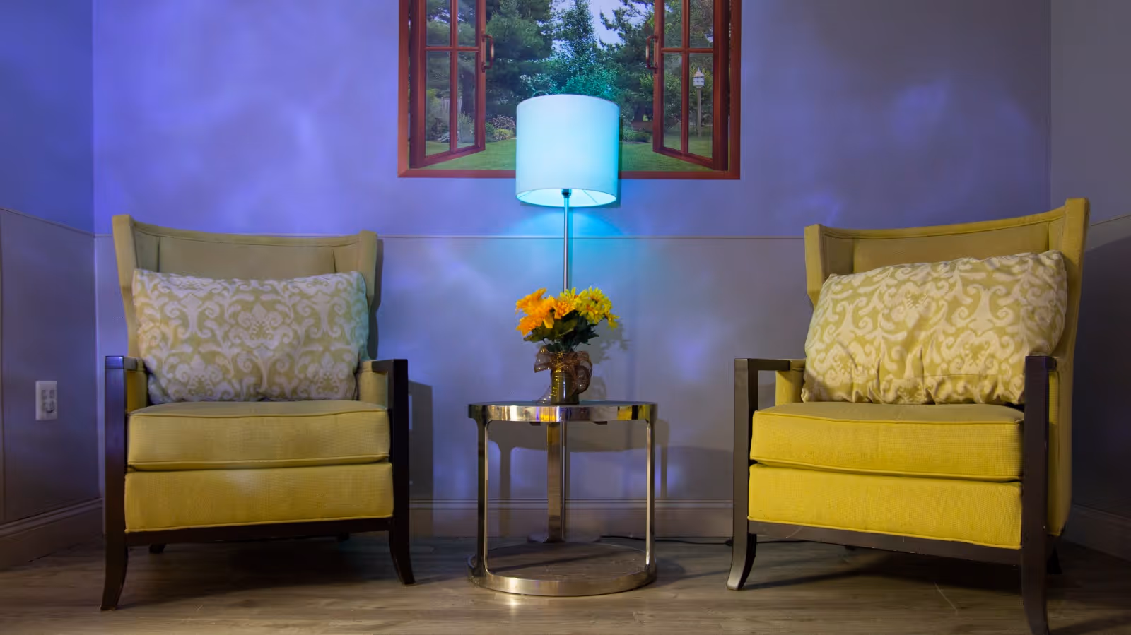 Two yellow armchairs with patterned cushions are placed on either side of a small round glass table with a vase of yellow flowers. Behind the table is a floor lamp with a blue shade, and above it is a wall decoration resembling an open window showing a green outdoor scene.