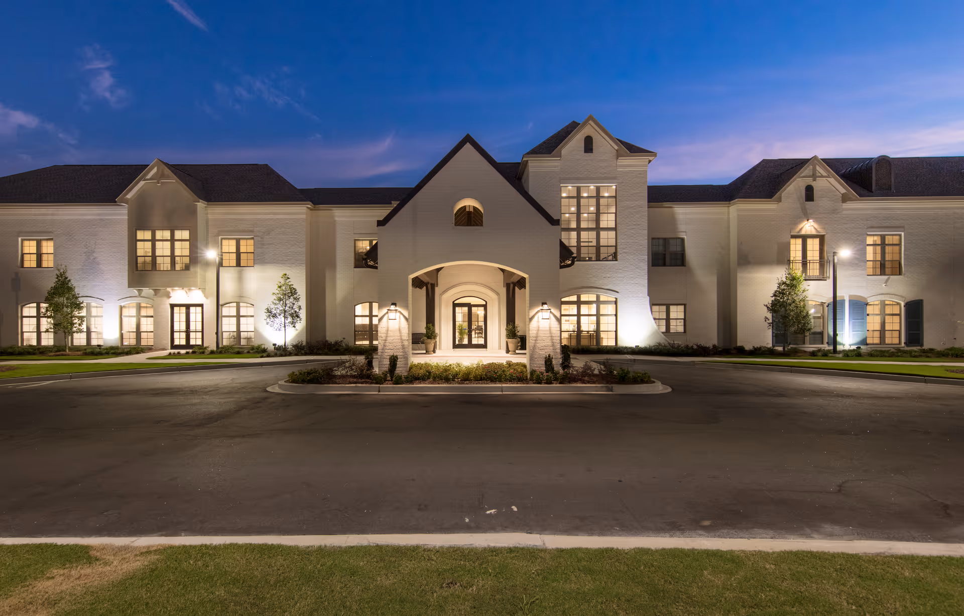 Front exterior view of The Blake at Carnes Crossroads building at dusk, showing a large, well-lit two-story white brick structure with multiple windows, a central entrance with an archway, and landscaped greenery around the driveway.