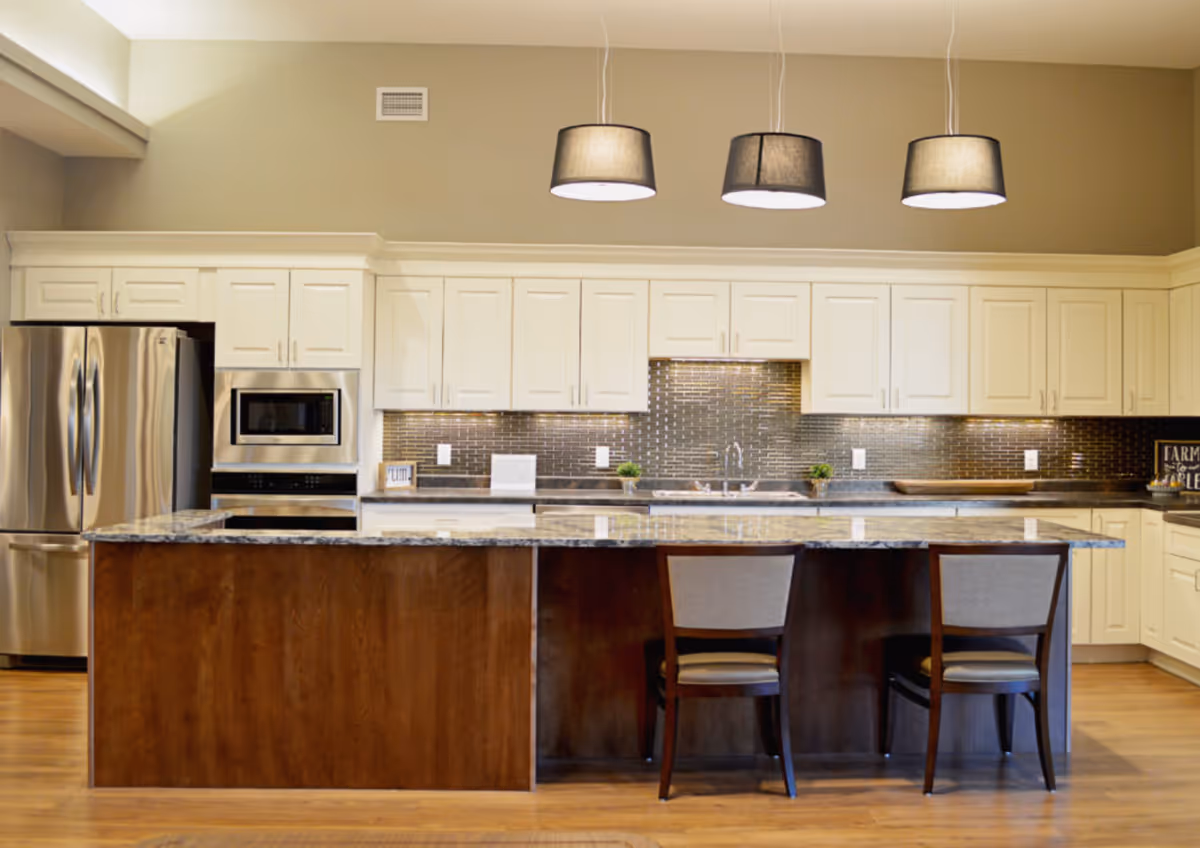Modern kitchen with white cabinets, stainless steel refrigerator and microwave, a large island with granite countertop, two chairs, and three pendant lights hanging from the ceiling.