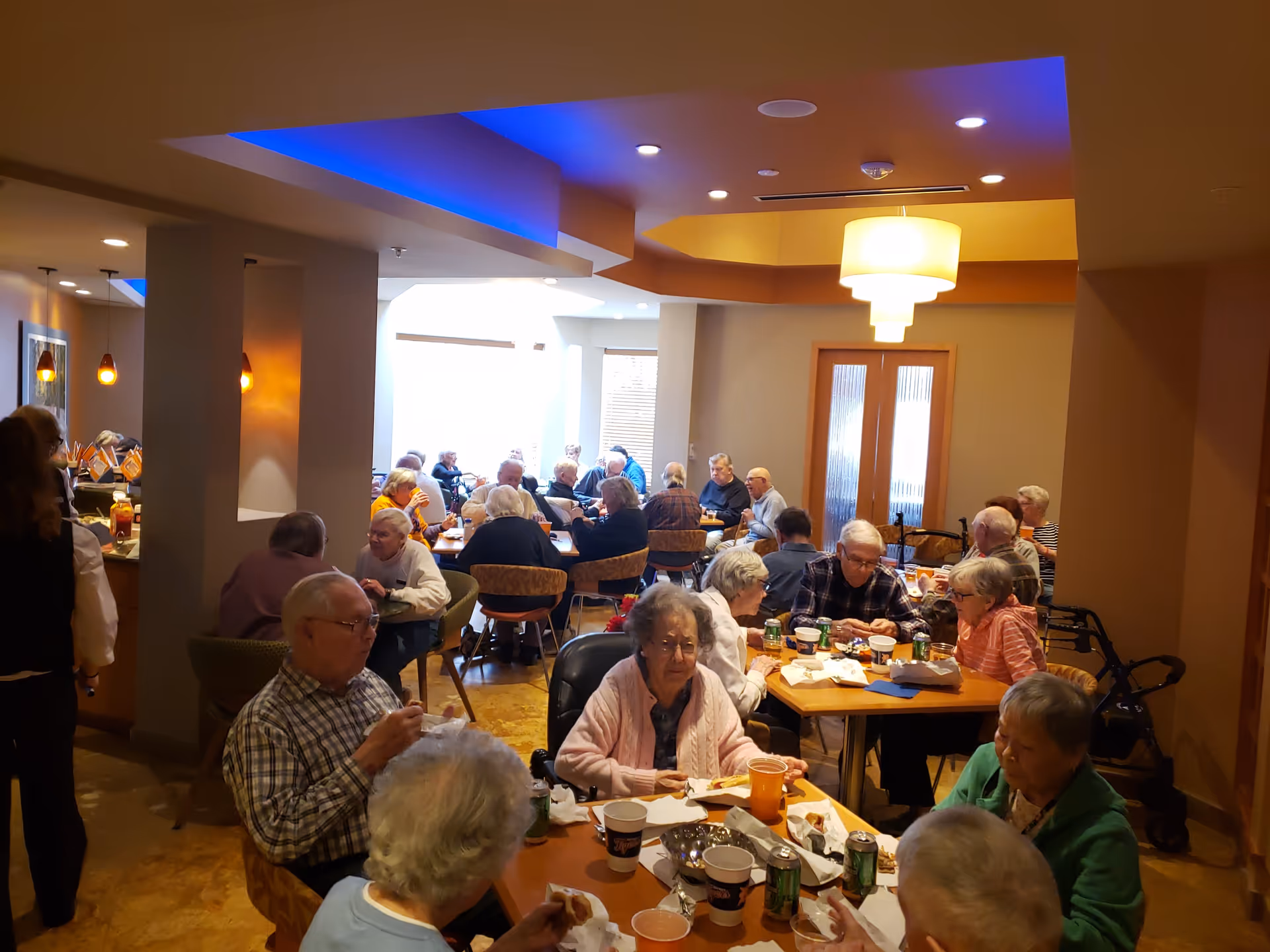 Elderly residents eating and socializing at multiple tables in a warm, well-lit communal dining room.