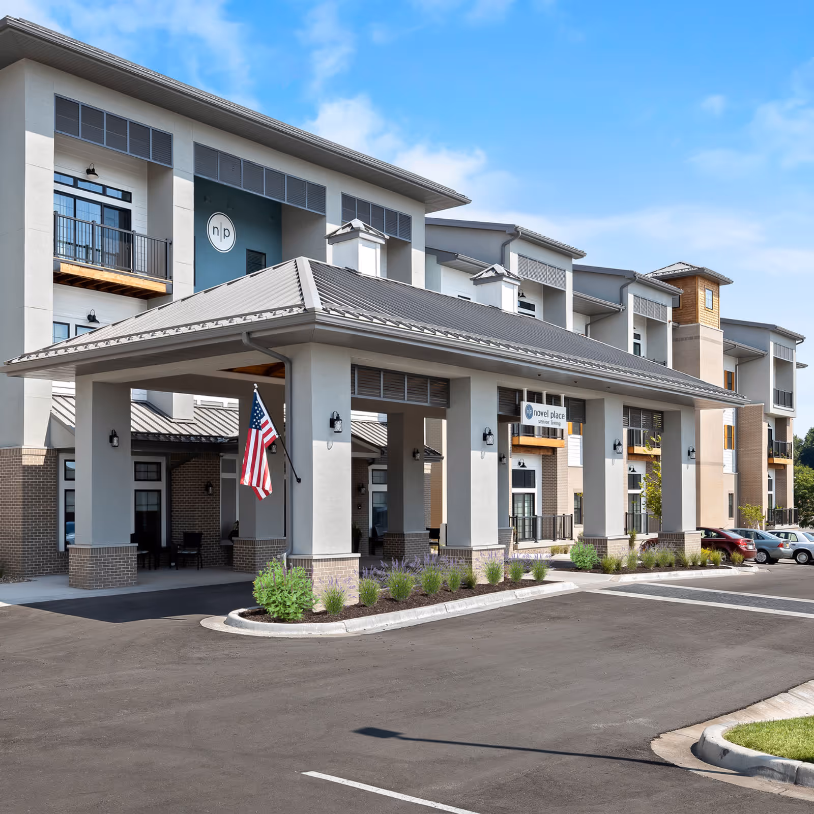 Exterior view of Novel Place Senior Living - Overland Park building with a covered entrance, American flag, multiple balconies, and parked cars under a clear blue sky.