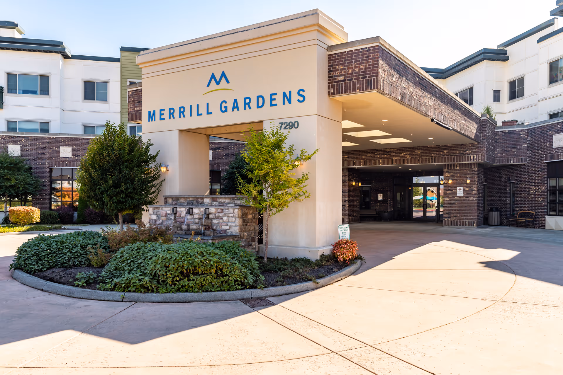 Entrance of Merrill Gardens at Tacoma senior living facility with a covered drop-off area, landscaped greenery, and the building's name prominently displayed on a large sign structure.
