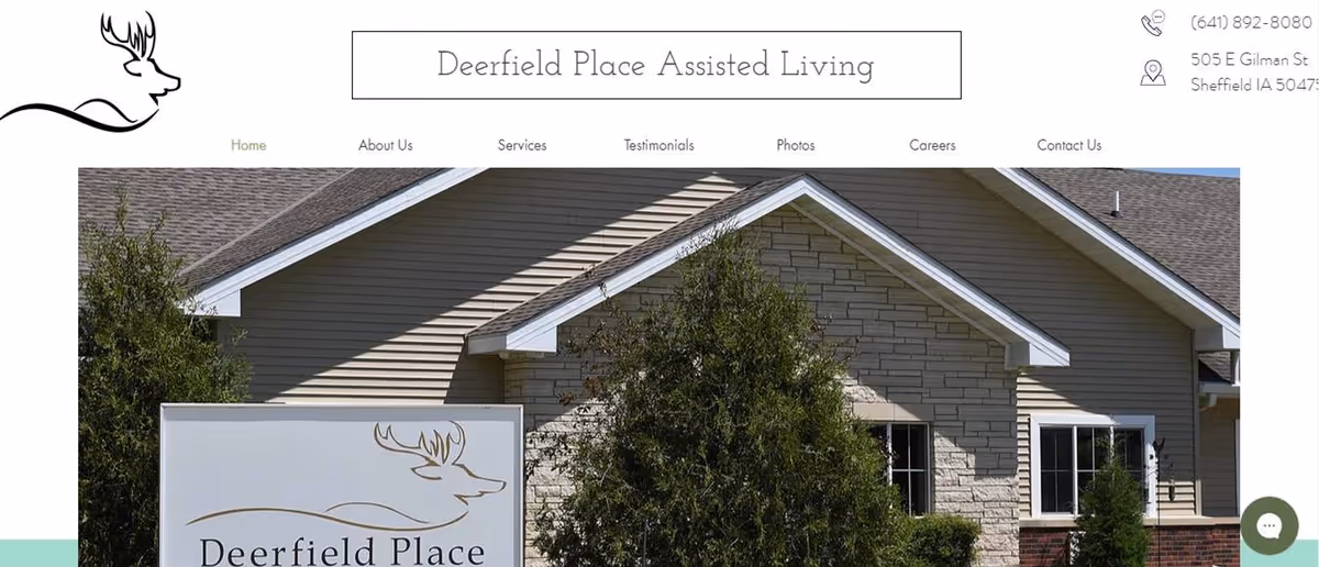 Exterior view of Deerfield Place Assisted Living building with beige siding and stone facade, partially obscured by green trees. A sign in front displays the Deerfield Place logo featuring a deer head outline and the facility name.