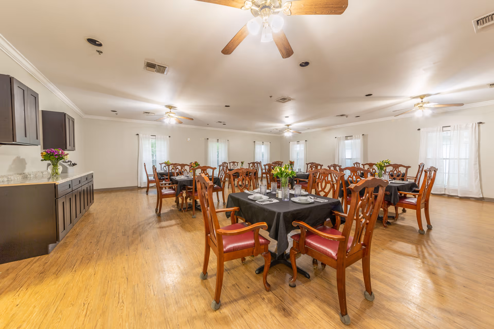 A spacious dining room with multiple wooden tables covered with black tablecloths, each set with plates, bowls, utensils, and glasses. Wooden chairs with red cushions surround the tables. The room has light-colored walls, several windows with white curtains, ceiling fans with lights, and a wooden floor. A counter with cabinets and a vase of flowers is visible on the left side.