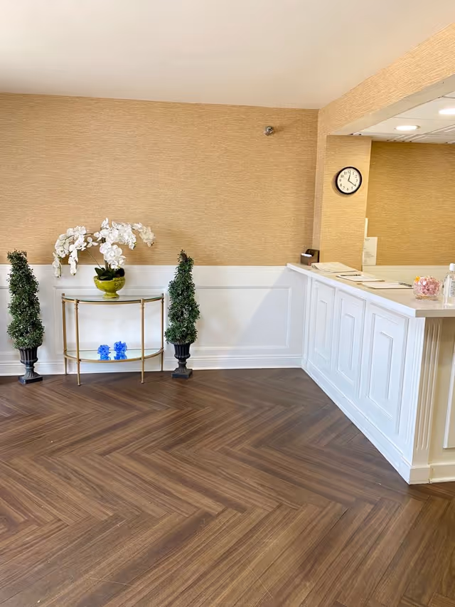 Reception area with a white counter on the right, a clock on the wall showing 11:10, and a small decorative table against a beige wall with two potted topiary plants and a white orchid in a green vase.