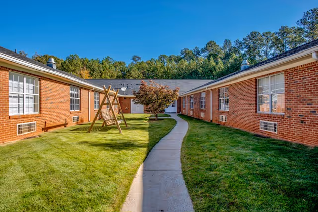 A courtyard area between two single-story brick buildings with multiple windows. There is a concrete pathway running through the center of the grassy area, which includes a wooden swing set and a small tree. Trees and a clear blue sky are visible in the background.