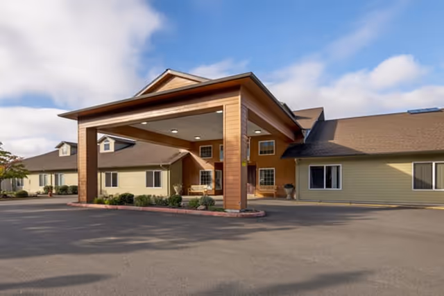 Front exterior of a single-story senior living building with a covered porte-cochere entrance, paved driveway, and blue sky.